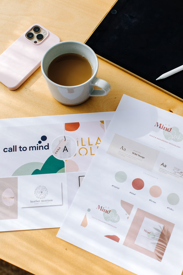 A phone, a mug of coffee, an iPad with a stylus, and printed branding materials featuring logos and design elements sit on a wooden surface—perfectly illustrating the essentials for service-based businesses specialising in graphic & web design.
